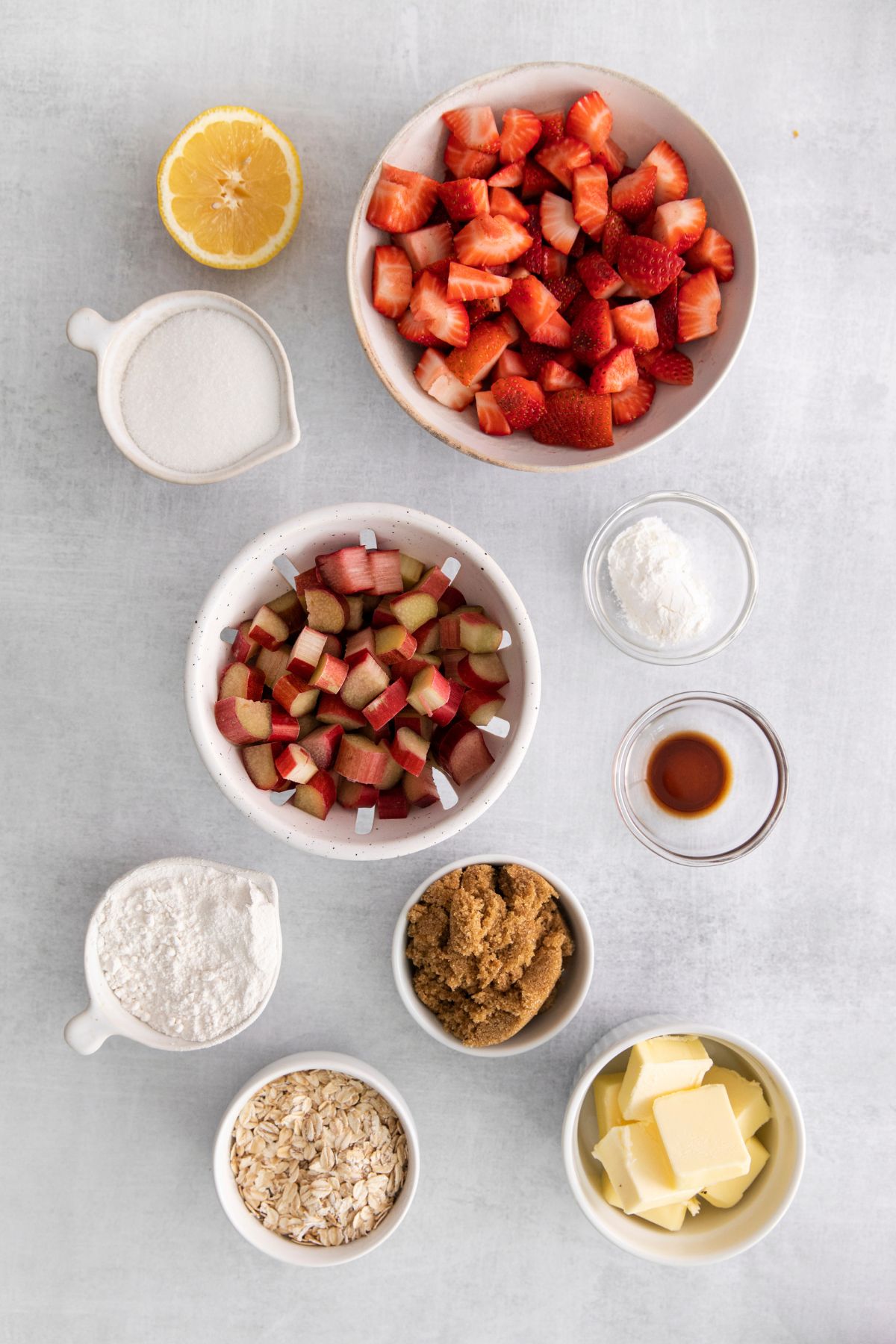 ingredients in bowls on white counter: half lemon, cut strawberries, cut rhubarb, sugar, flour, cornstarch, vanilla, butter, oats