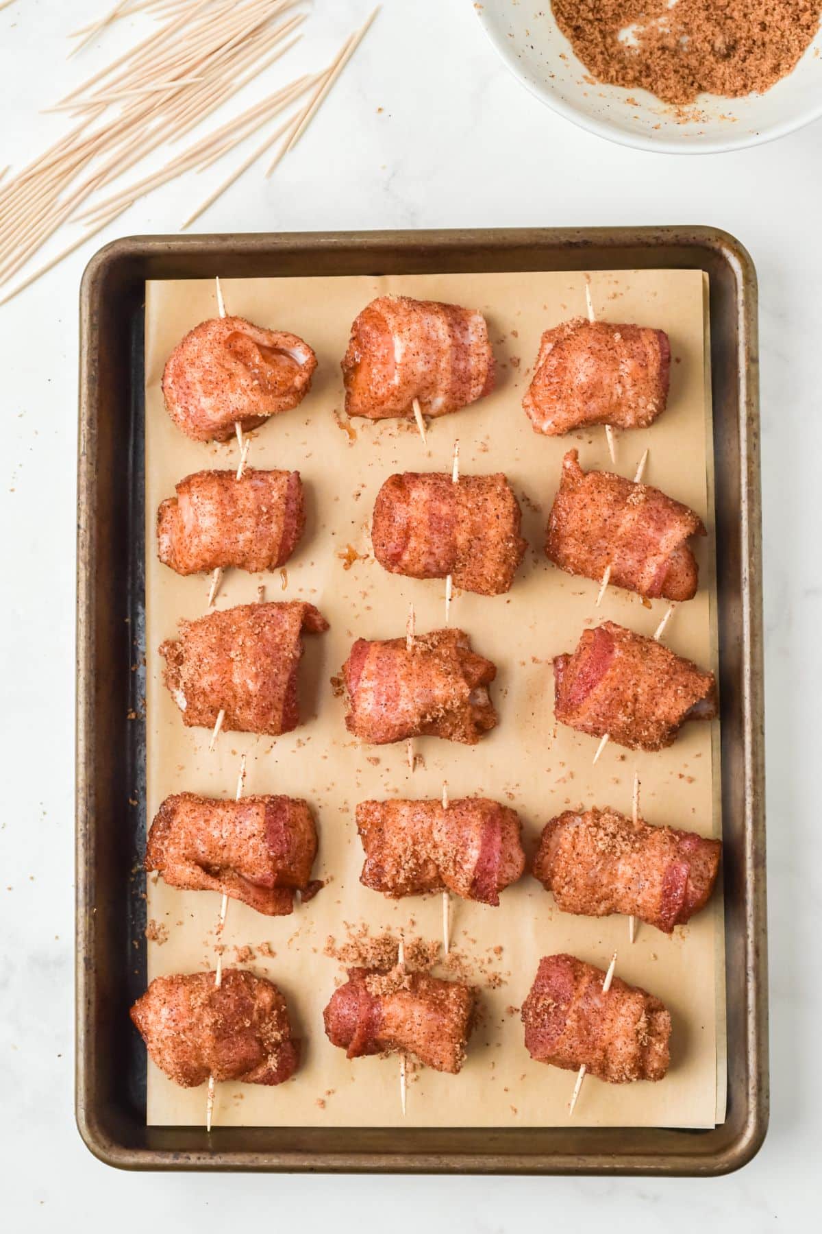 chicken wrapped in bacon with seasonings and a toothpick through each piece on a baking sheet