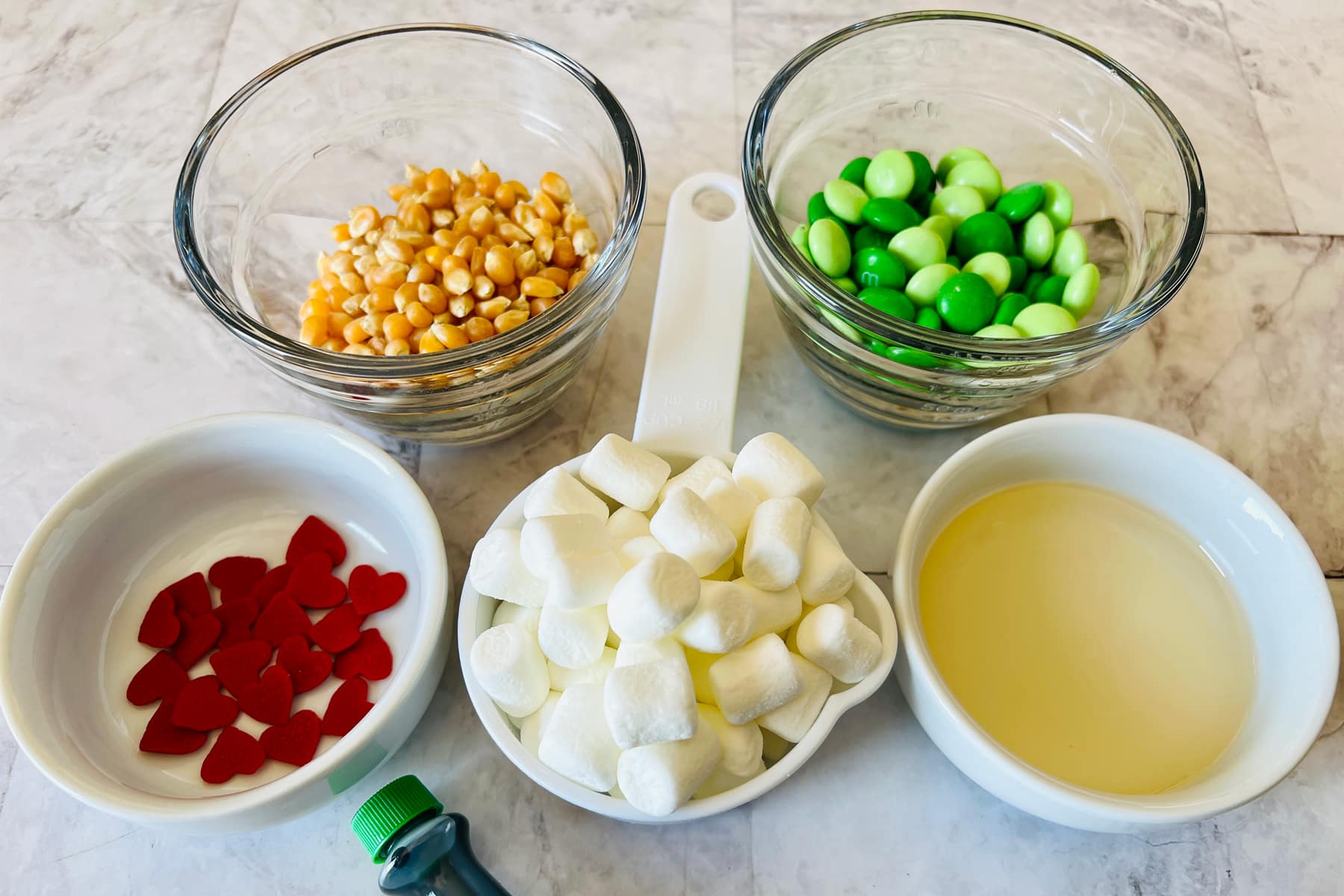 Grinch popcorn ingredients include ½ cup popcorn kernels, 2-3 tablespoons vegetable oil, Pinch of salt, Green regular and oil based food coloring, Green M&M candies, Mini marshmallows, Red decorator candy hearts