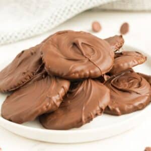 A white plate holds a pile of round Ritz Crackers Thin Mints, each coated in a smooth layer of chocolate, arranged on a light surface.