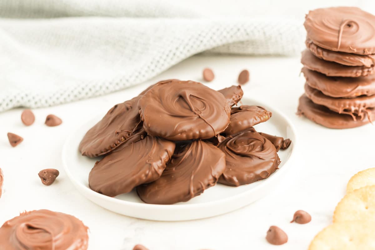 A white plate with a stack of chocolate-covered cookies made from Ritz Crackers Thin Mints, surrounded by chocolate chips and more cookies on a white surface.