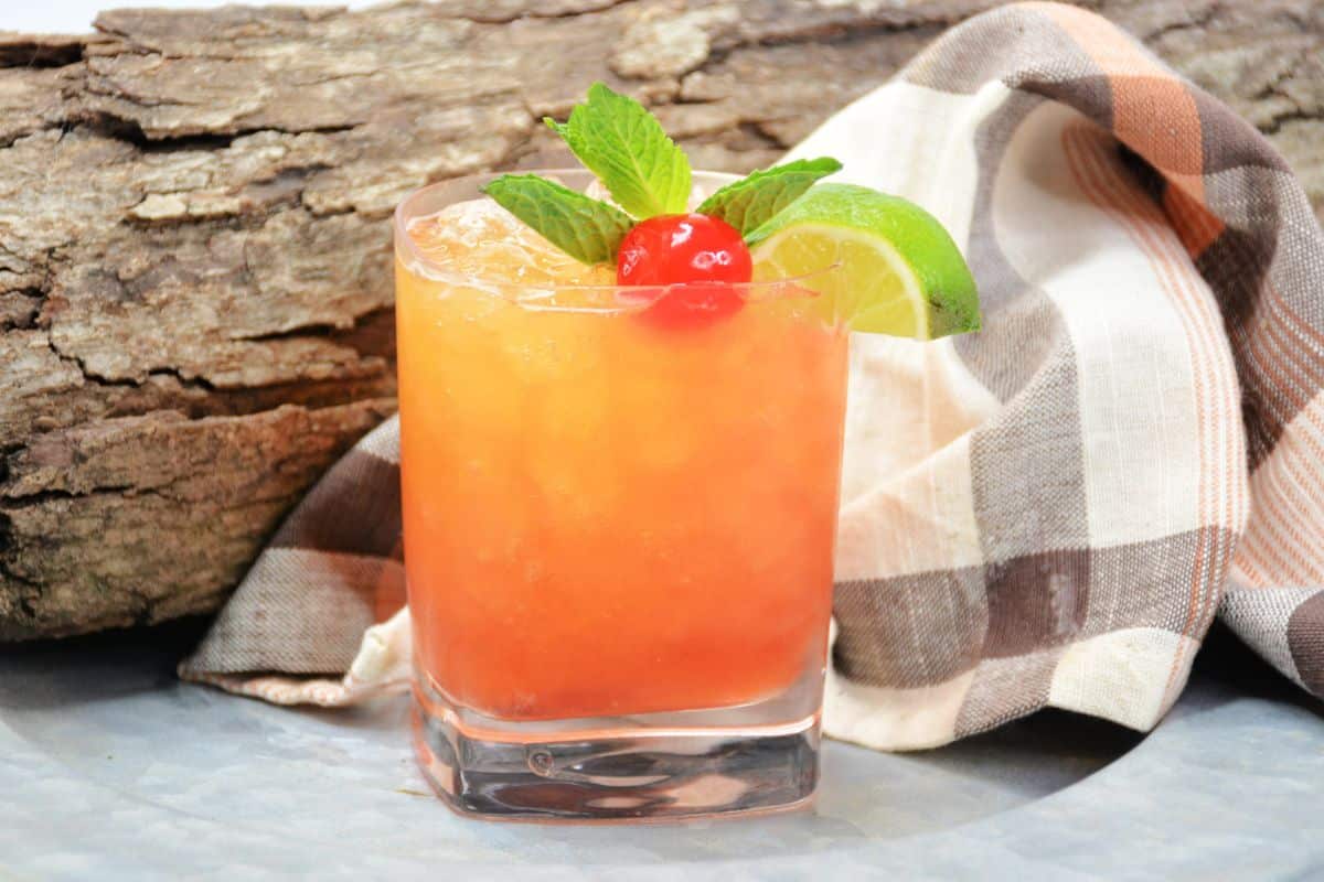 orange drink in clear cocktail glass with cherry and lime next to wooden log