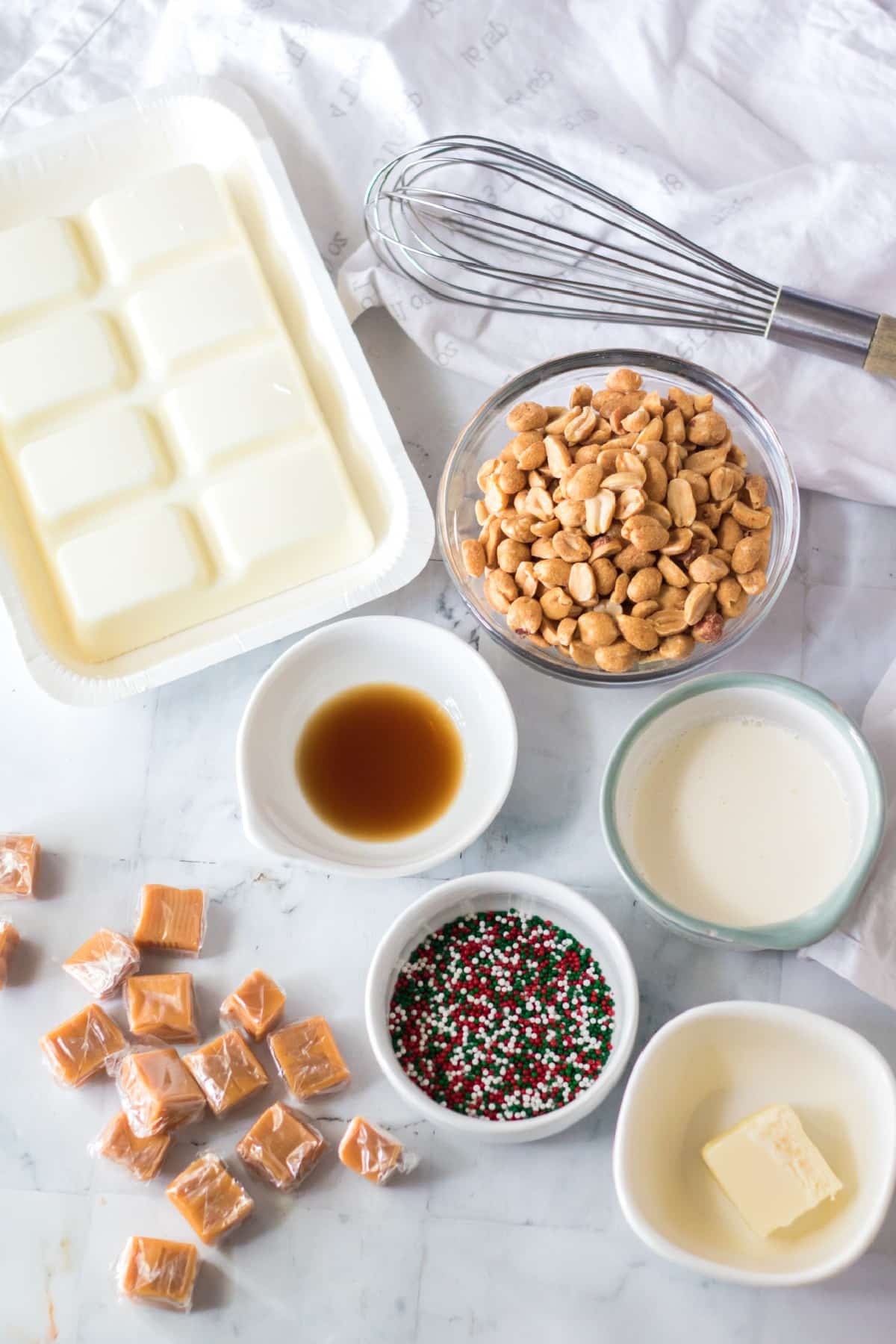Polar Bear Claws ingredients on a white counter. These are the following: caramel squares, whipping cream, butter, vanilla extract, salted peanuts, almond bark, and sprinkles