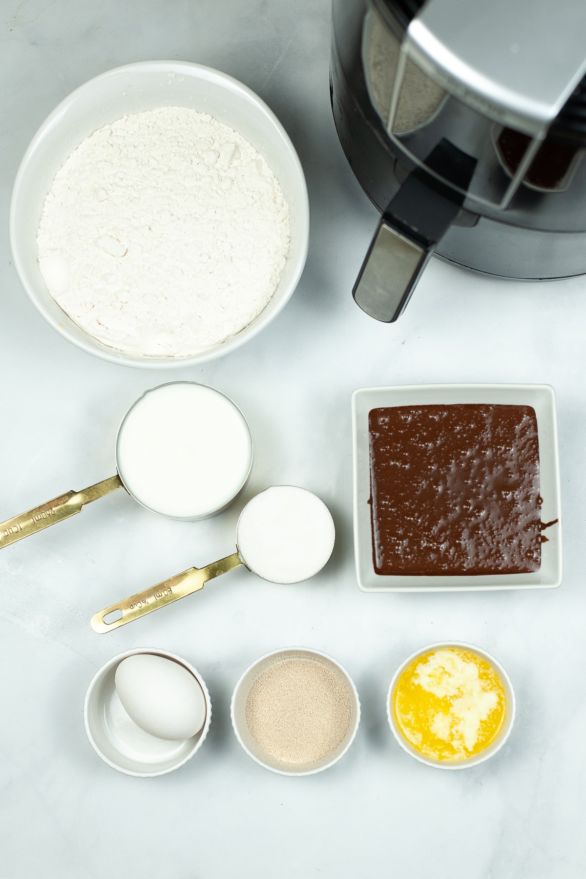 Ingredients for Chocolate Glazed Air Fryer Donuts on a white counter. These include milk, sugar, yeast, flour, melted butter, egg and chocolate frosting.