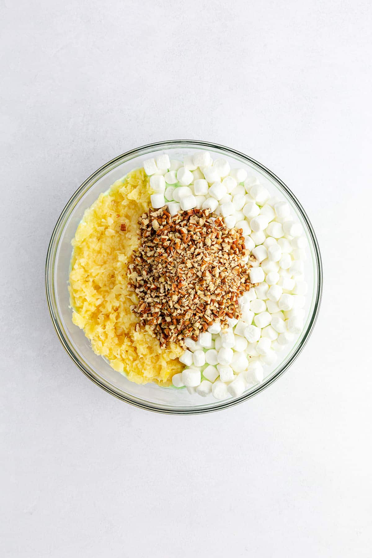 White mallows, chopped pecans, and crushed pineapple are arranged artfully in a glass bowl