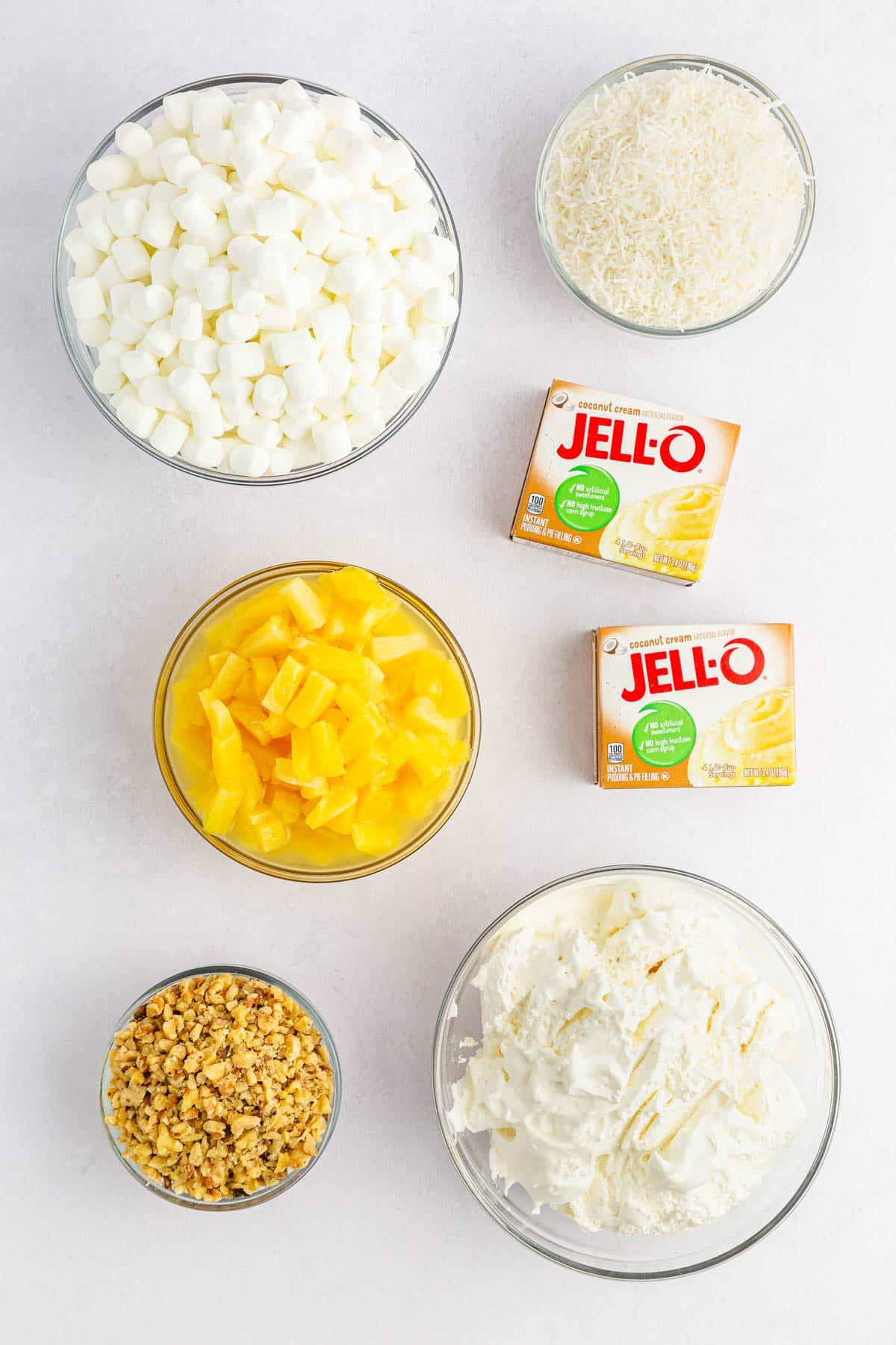 marshmallows, coconut, jello, whipped topping, pineapple chunks, and nuts on counter in bowls