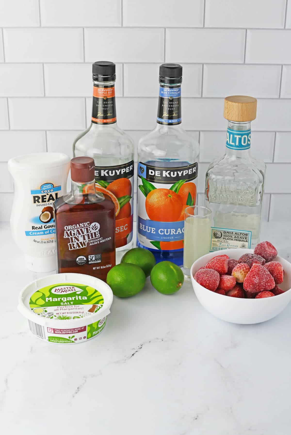 liqueur bottles with cream of coconut, agave and lines, margarita salt and frozen strawberries on counter