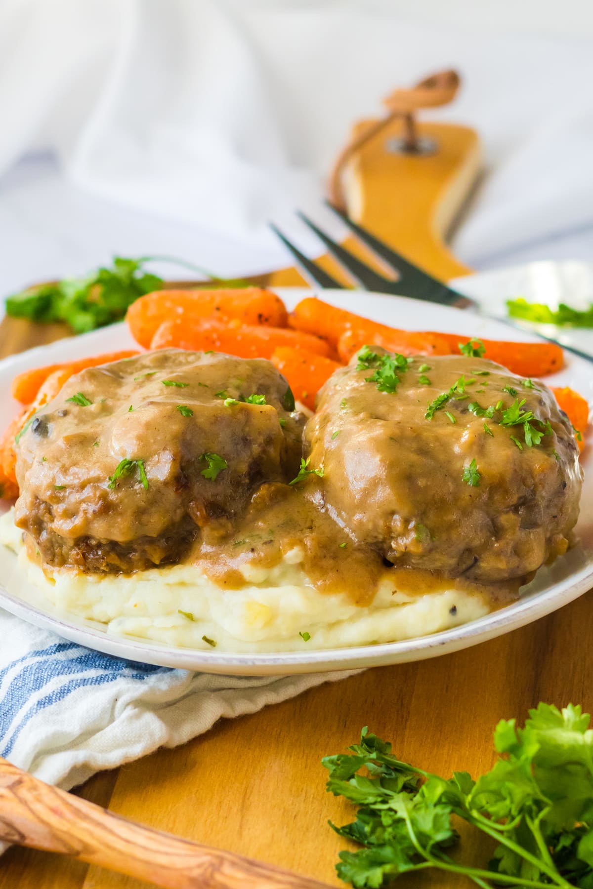 A serving of Slow Cooker Smothered Hamburgers in a white plate with veggies and spoon on the side.