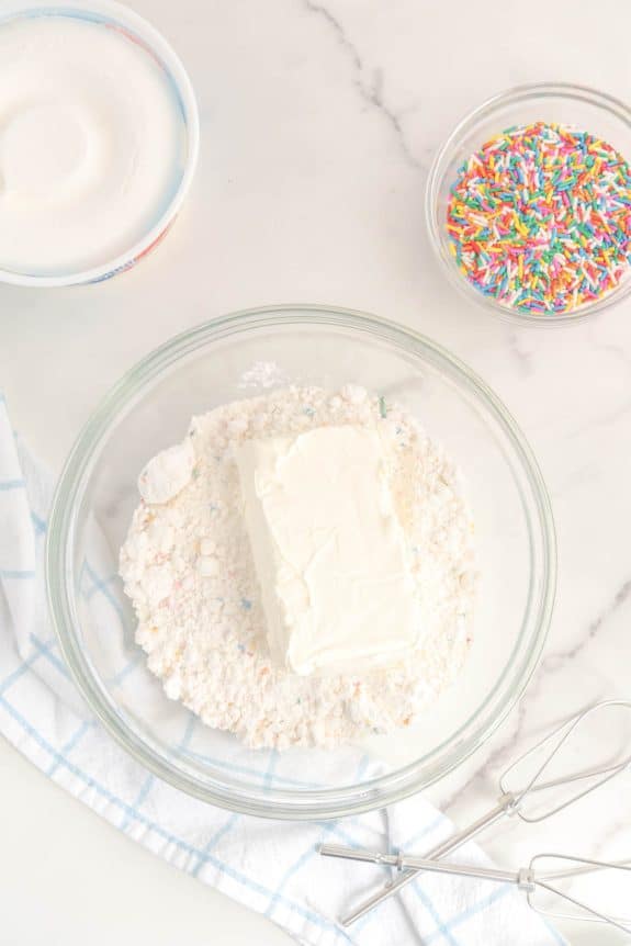 A bowl of icing, a whisk, and a bowl of sprinkles.
