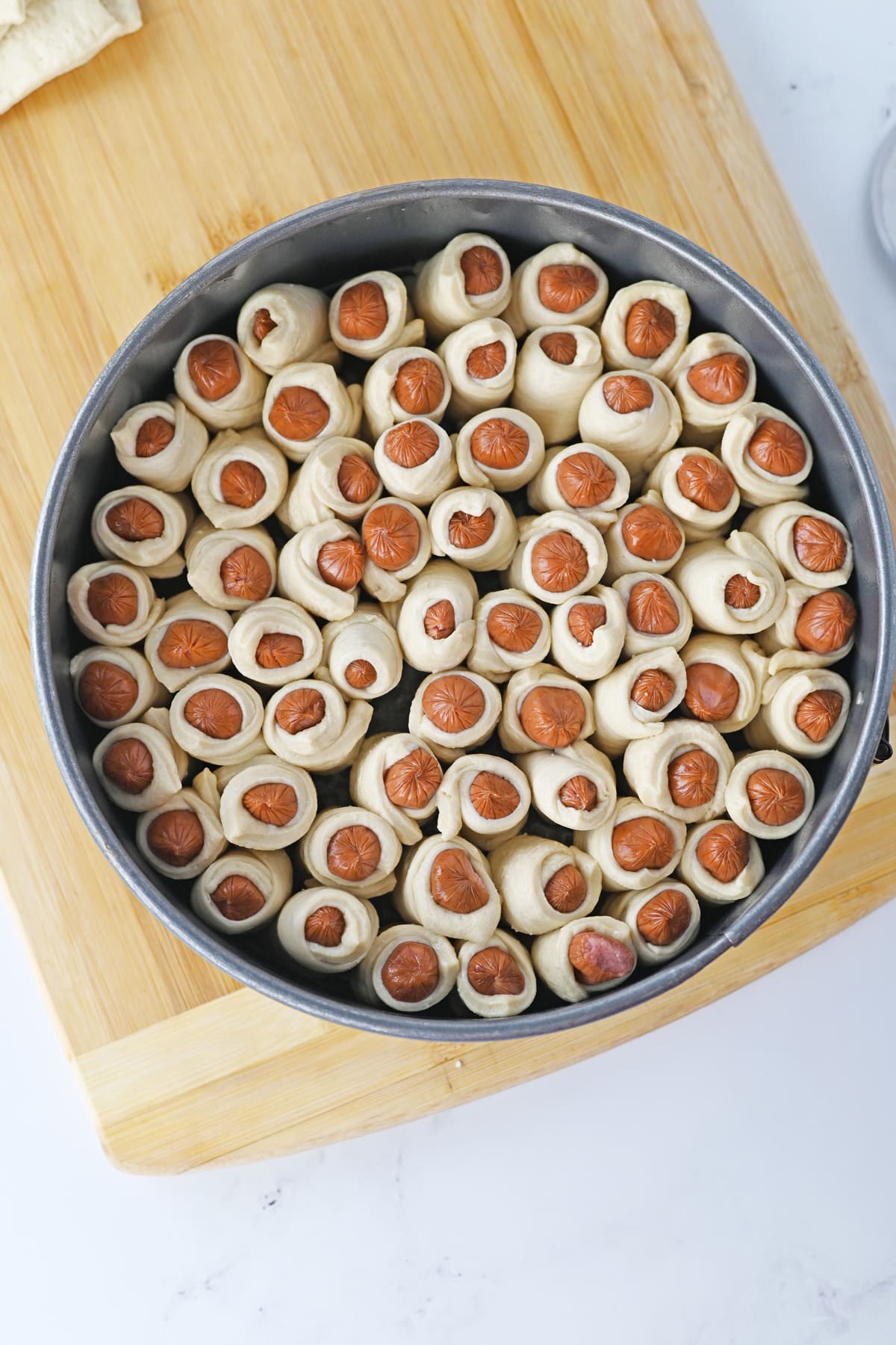 Next step in making Pigs in a Blanket is to prepare hot dog rolls in a pan on a cutting board.