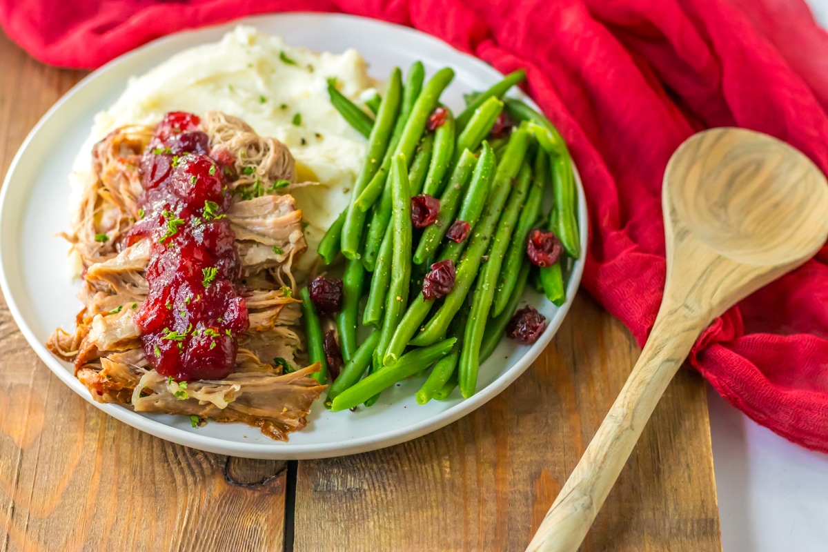 A plate of Slow Cooker Cranberry Pork with vegetables, linen and wooden spoon on the side.