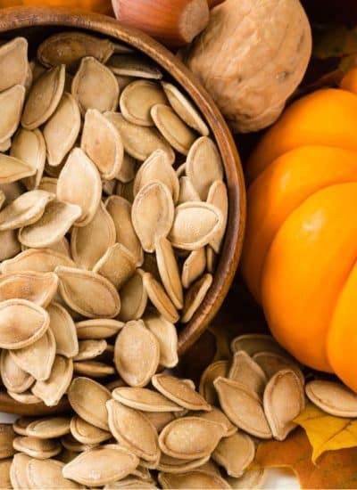 A bowl of saved pumpkin seeds, surrounded by leaves and pumpkins, awaits their use in a delicious meal.