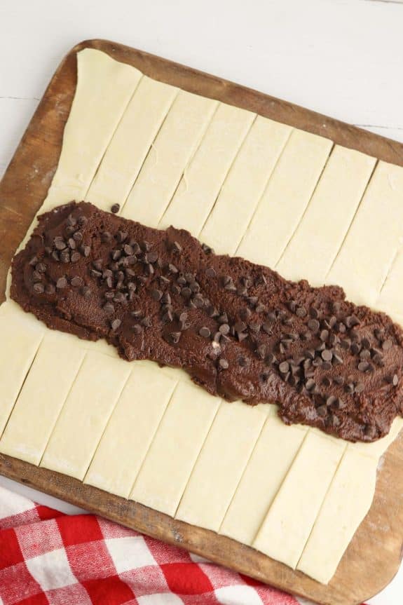 Chocolate chips and nutella on puffed pastry on a cutting board.