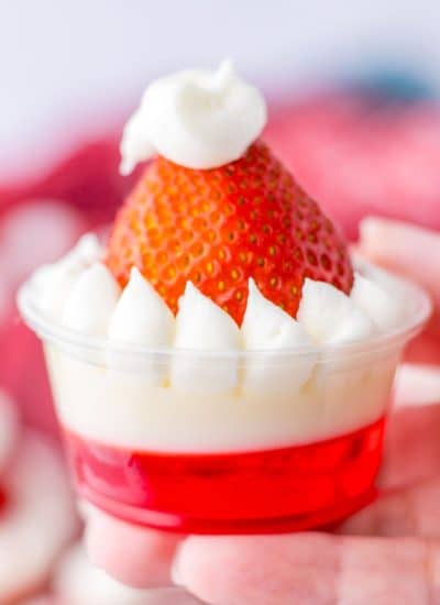 A person holding a strawberry jello dessert in a cup topped with a Santa hat.