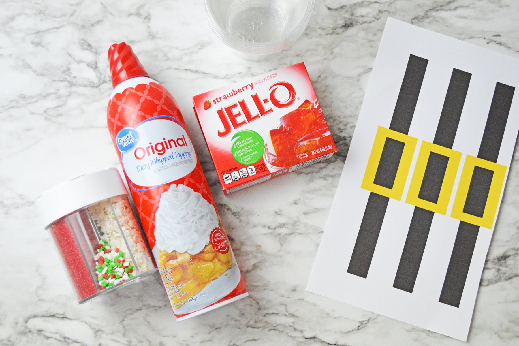 The ingredients for Santa Jello Cups for kids include strawberry jello, water, divided, Santa’s belt printable (printed with belts cut out), scotch tape, whipped cream for topping, Christmas sprinkles for topping (optional), and clear solo cups