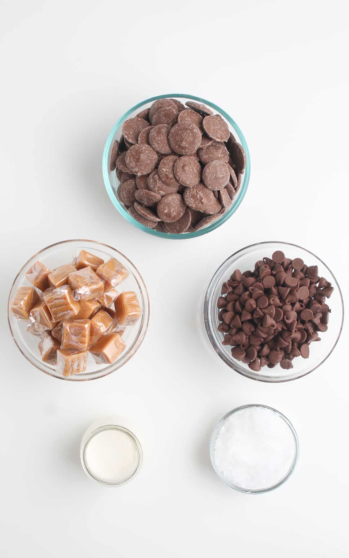 A bowl of chocolate chips, caramels, chocolate melting wafers, sugar and cream on white counter.