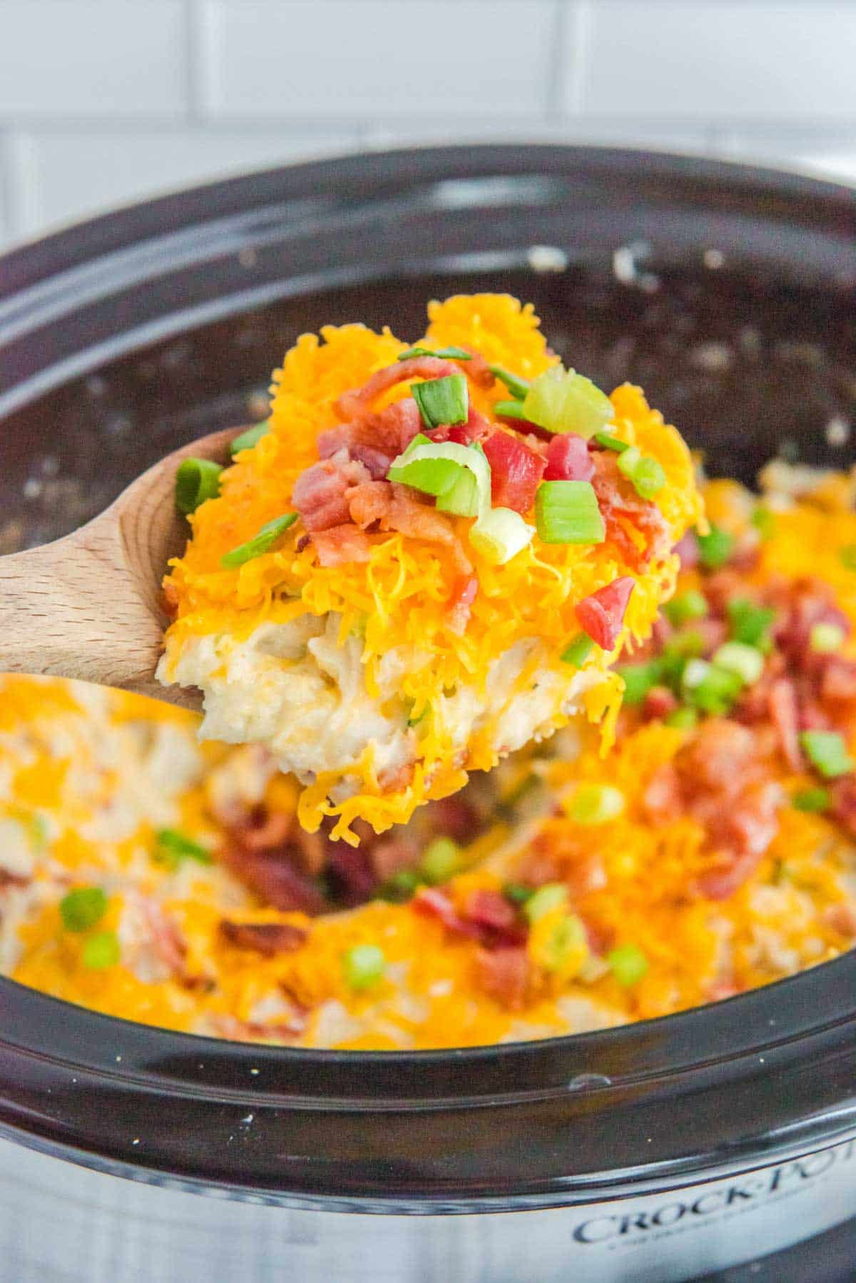 A wooden spoon is being used to scoop out a slow cooker loaded mashed potatoes casserole.