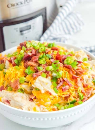 A bowl of slow cooker loaded mashed potatoes with cheese and bacon in front of it.
