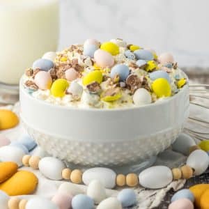 A white bowl filled with colorful Easter treats, including Cadbury eggs, and accompanied by a glass of milk.