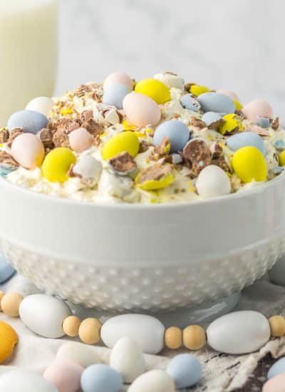 A white bowl filled with colorful Easter treats, including Cadbury eggs, and accompanied by a glass of milk.