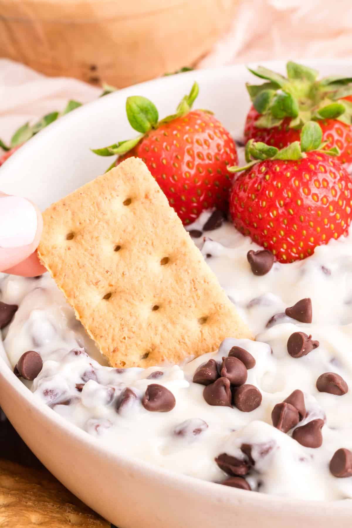 A person is dipping a cracker into a bowl of Cannoli Dip with strawberries and chocolate chips.