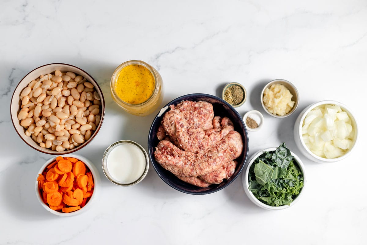 Ingredients for Kale Sausage White Bean Soup in a bowl. This include the following: Italian pork sausage, onion, minced garlic, carrots, canned cannellini beans, chicken broth, dried oregano, fresh kale, milk, salt and black pepper.