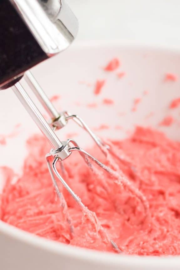 Pink frosting being mixed with a mixer in a white bowl.