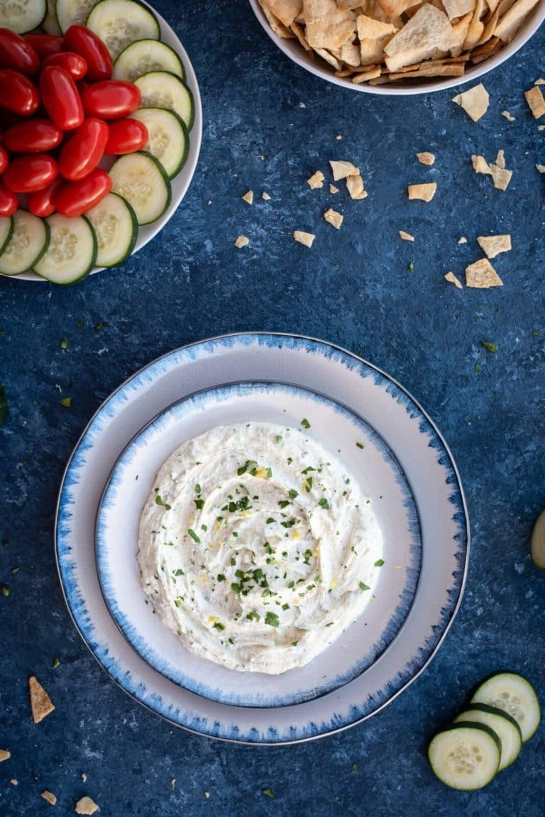 A bowl of hummus with cucumbers and tomatoes on a blue background.