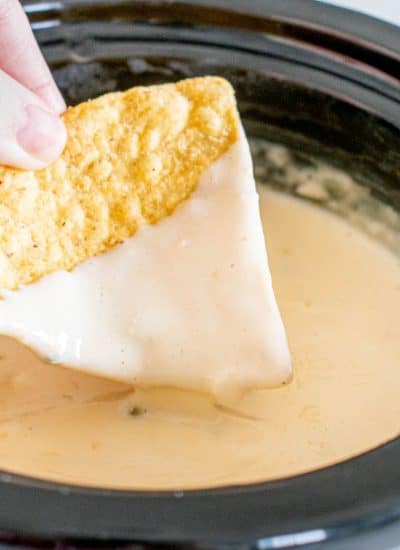 A person dipping a tortilla chip into a crockpot of white queso dip.