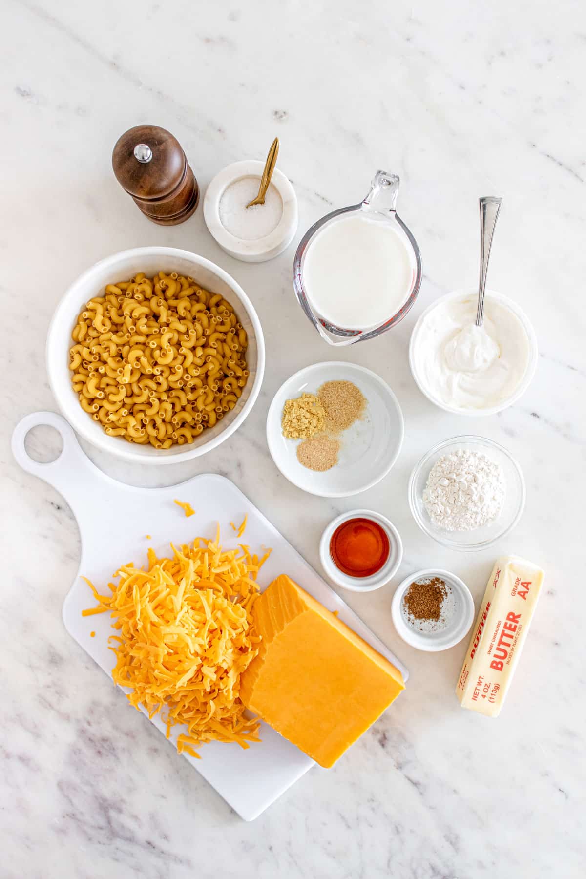 Ingredients for Stovetop Mac and Cheese on a white marble counter include elbow macaroni, butter, all-purpose flour, whole milk, sour cream, garlic powder, mustard powder and ground nutmeg, hot sauce, and sharp cheddar cheese.