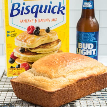 A freshly baked loaf of *Bisquick beer bread* on a cooling rack with a bisquick box and a bottle of Bud Light in the background.