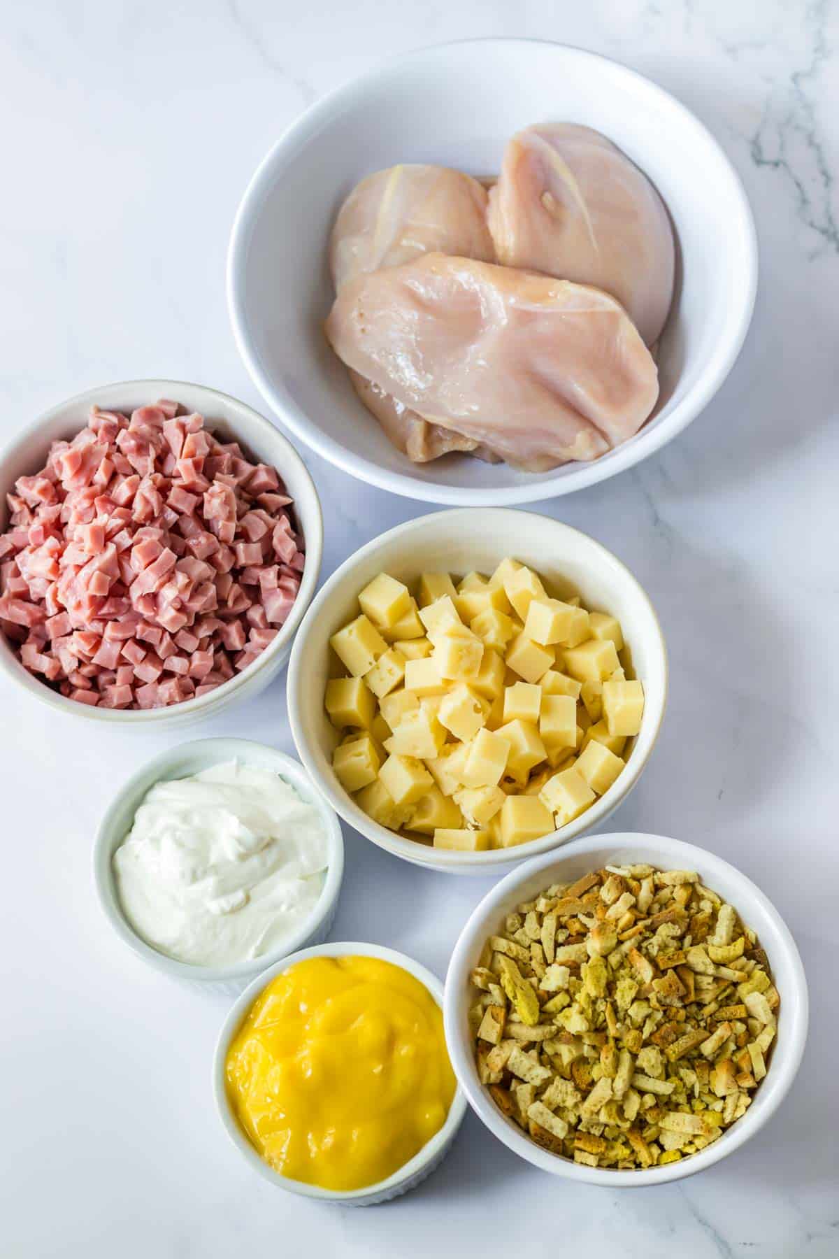 Various ingredients in bowls including chicken breasts, diced ham, cubed cheese, stuffing mix, sour cream, and mustard ready for a recipe preparation.