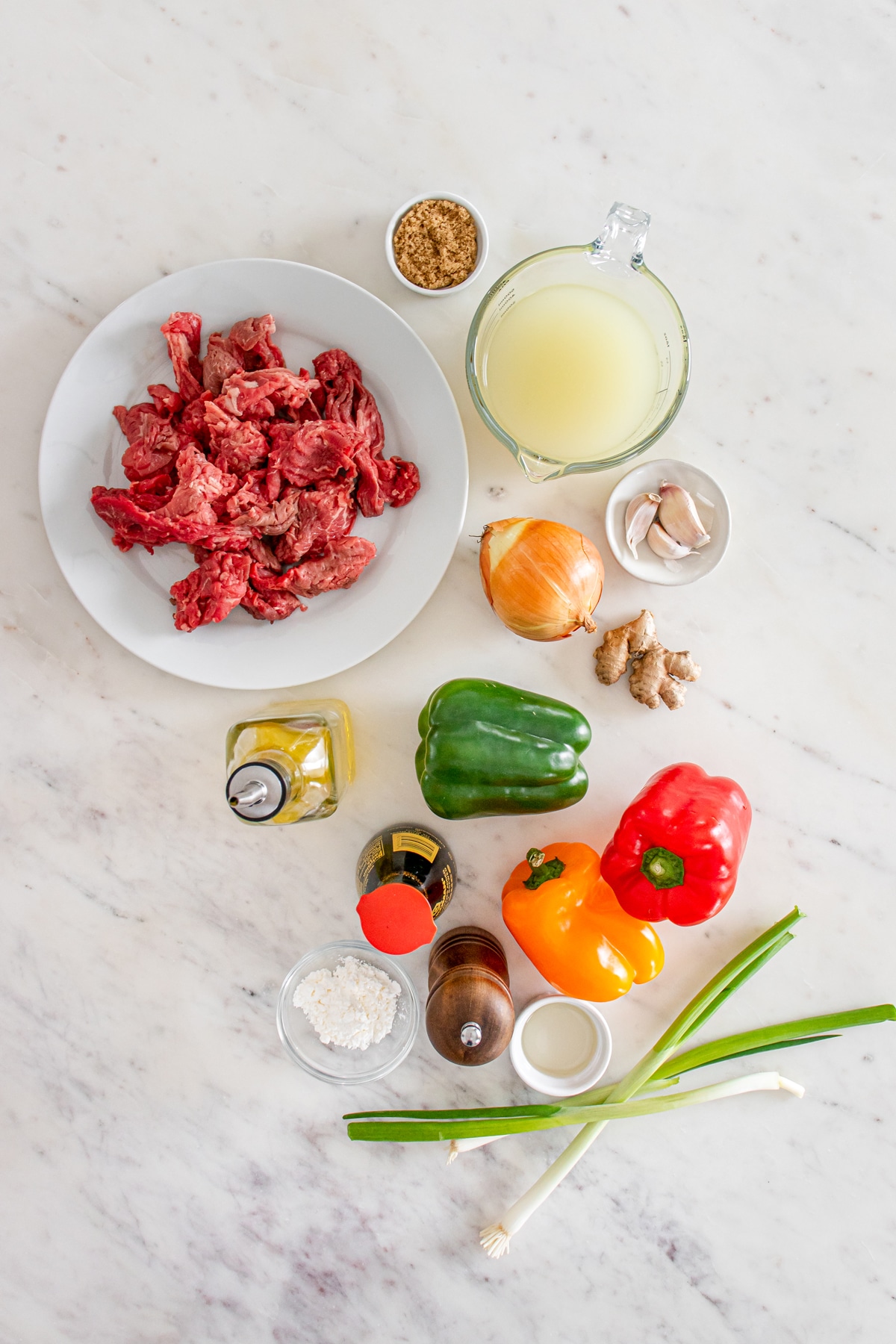 Assorted ingredients for cooking, including beef, bell peppers, onions, and spices, arranged neatly on a marble surface.