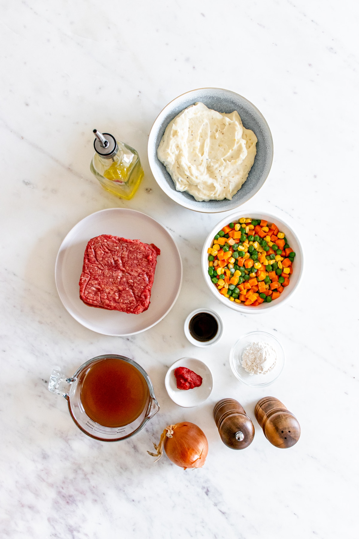 Ingredients for Shepherd's Pie on a white counter. These include: beef, mashed potatoes, olive oil, yellow onion, lean ground beef, all-purpose flour, beef broth, frozen mixed veggies, tomato paste and Worcestershire sauce, salt and pepper, and fresh parshley.