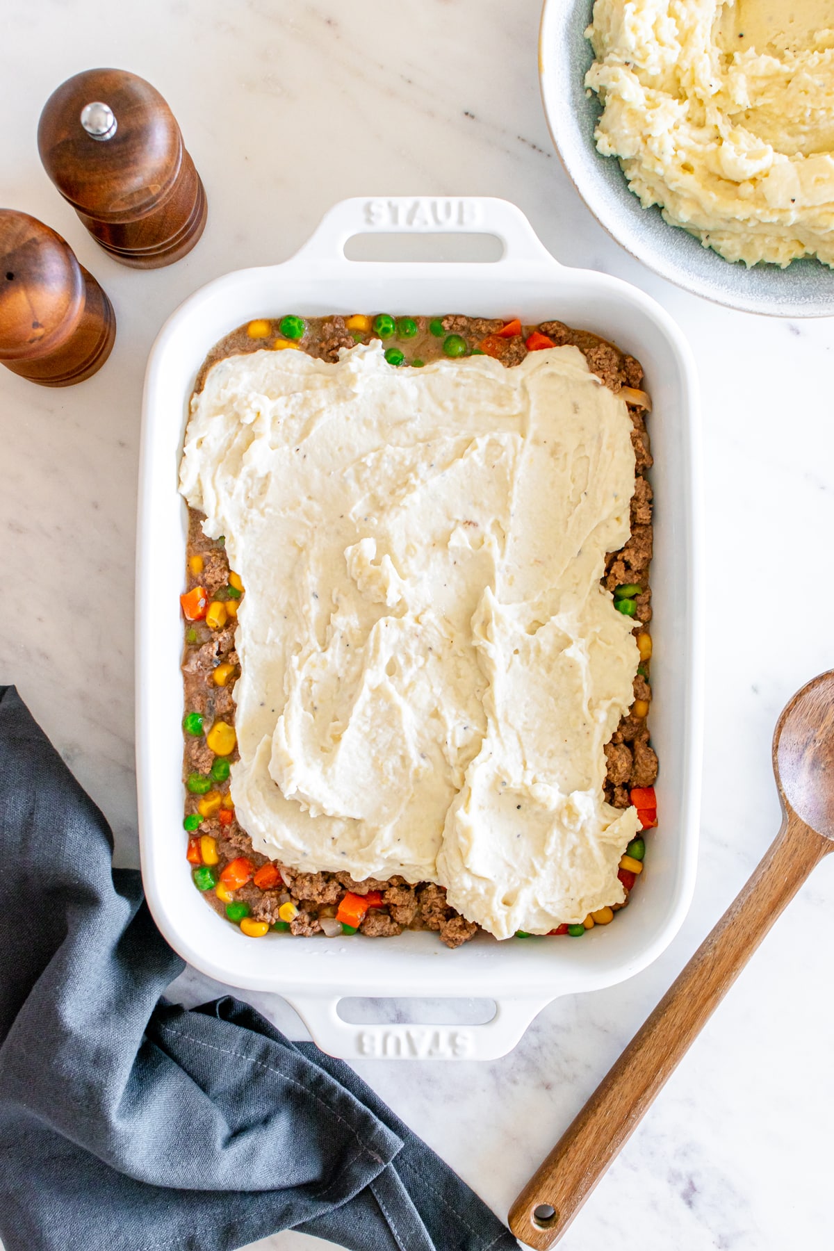 A shepherd's pie in a white baking dish, before being baked, with mashed potatoes on top of the meat and vegetable layer, next to wooden salt and pepper mills and a bowl of extra mashed potatoes.