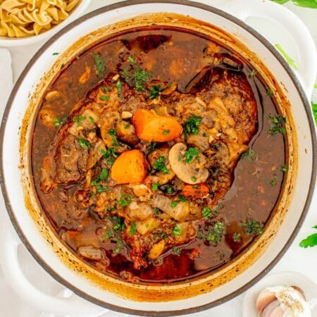 A pot of hearty beef stew with carrots, mushrooms, and herbs, viewed from above on a white tablecloth with scattered garlic cloves and pasta in the background.