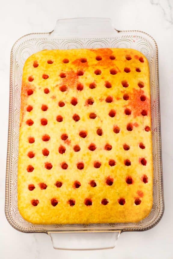 A freshly baked golden sponge cake with red jam-filled holes in a rectangular glass dish on a marble countertop.