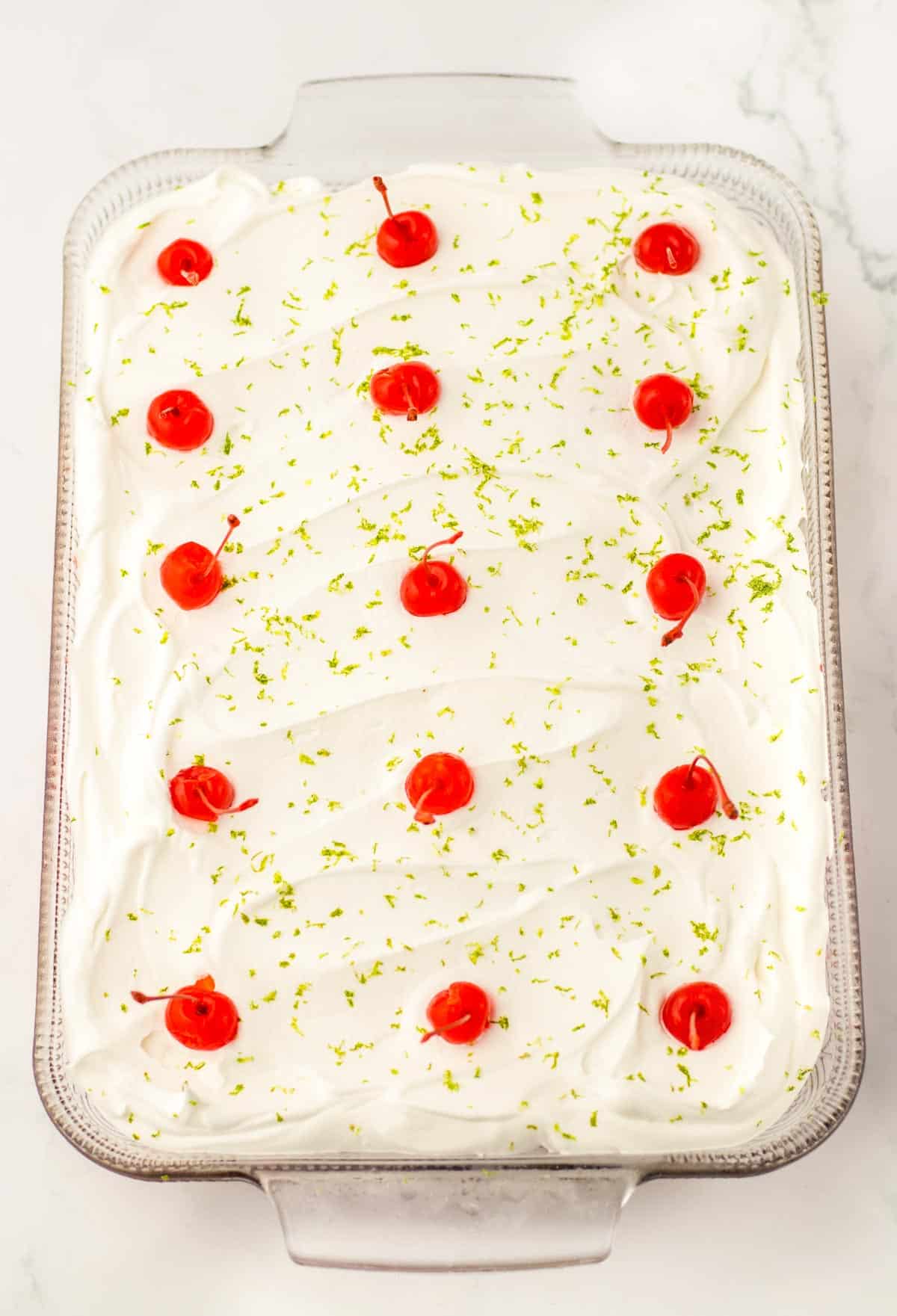 A tray of lime sheet cake topped with white frosting, red cherries, and sprinkled lime zest on a marble surface.