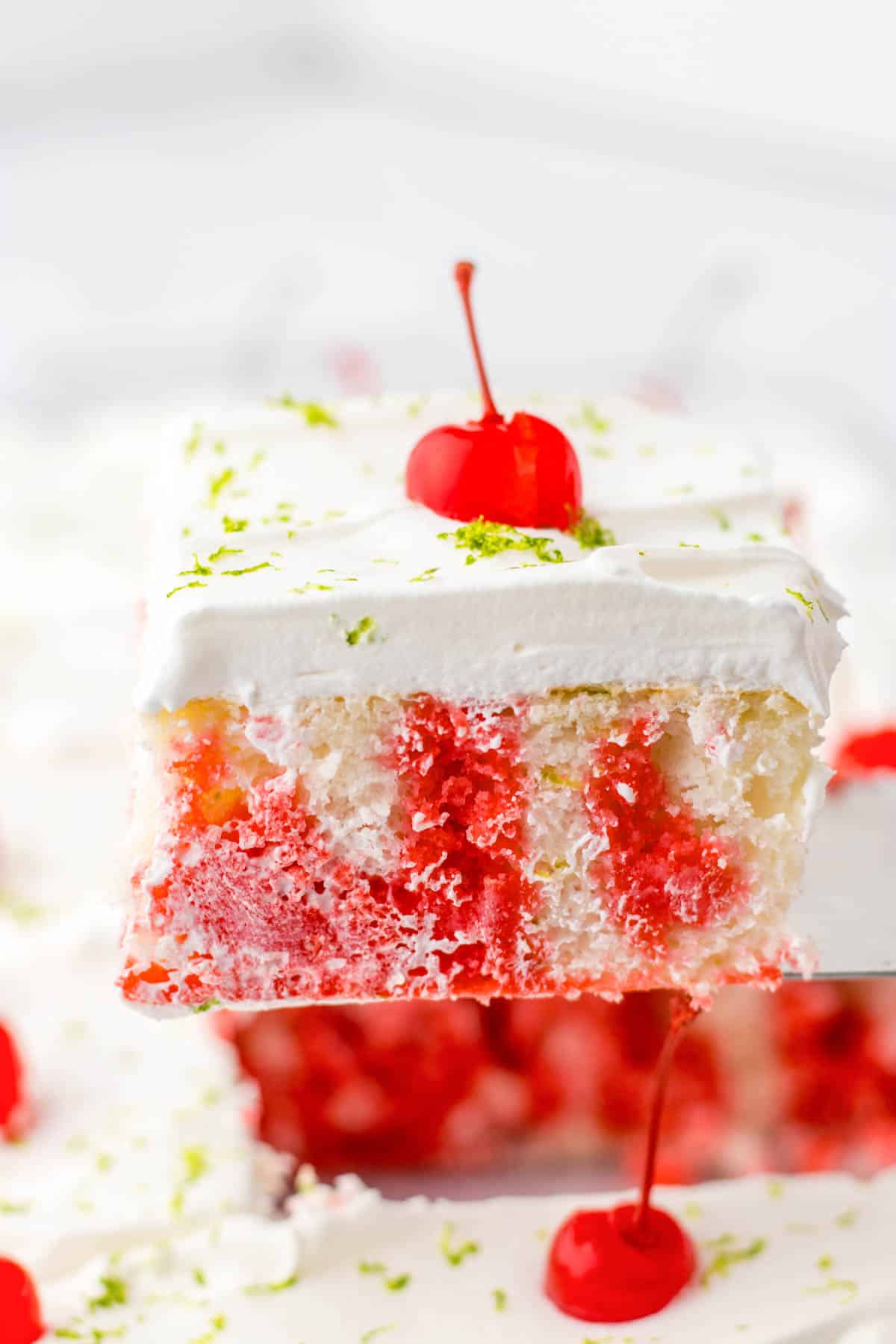 A slice of cherry lime poke cake topped with whipped cream and a cherry, garnished with lime zest.