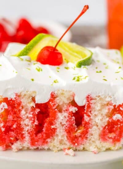 A slice of cherry poke cake with vibrant red layers, topped with white frosting, lime slices, and a cherry on a white plate.