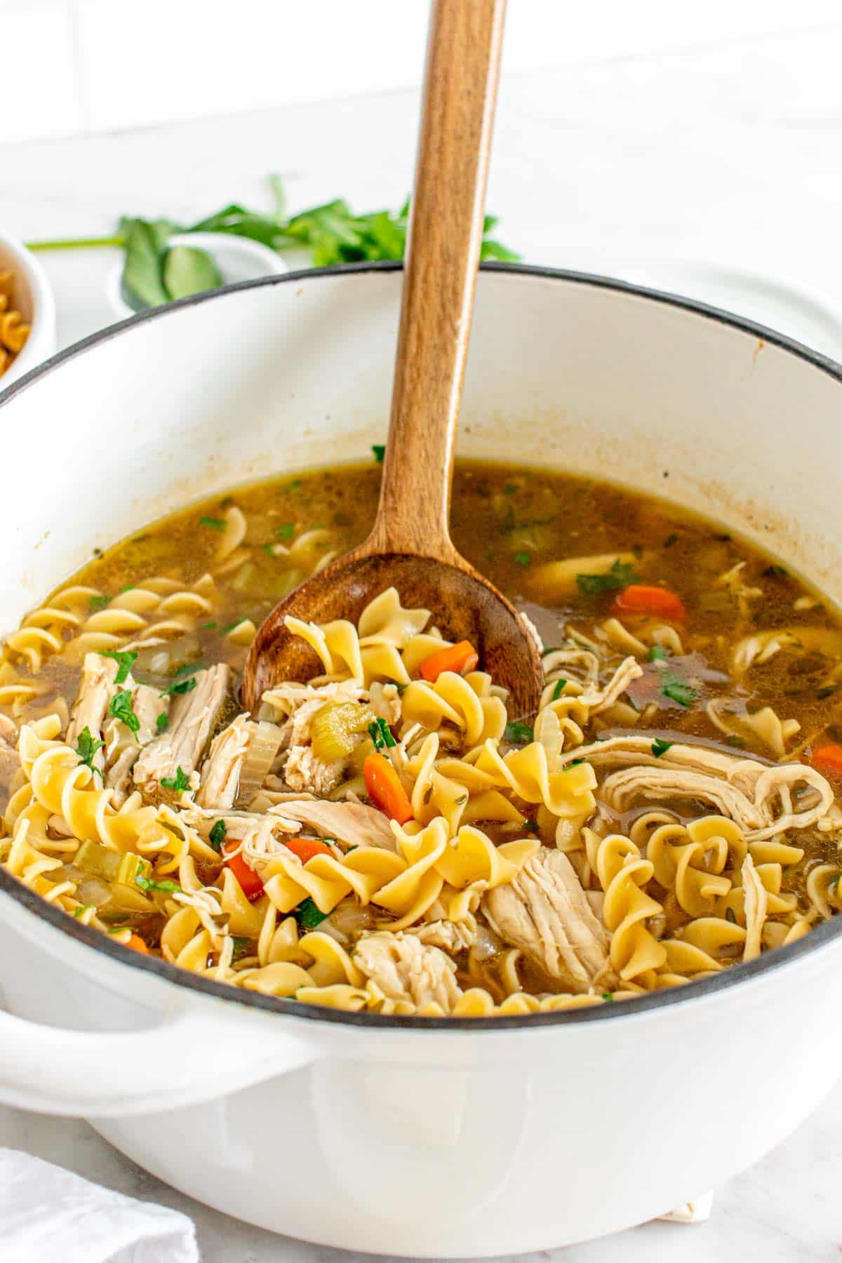 A wooden spoon lifting noodles from a pot of chicken noodle soup, garnished with herbs.
