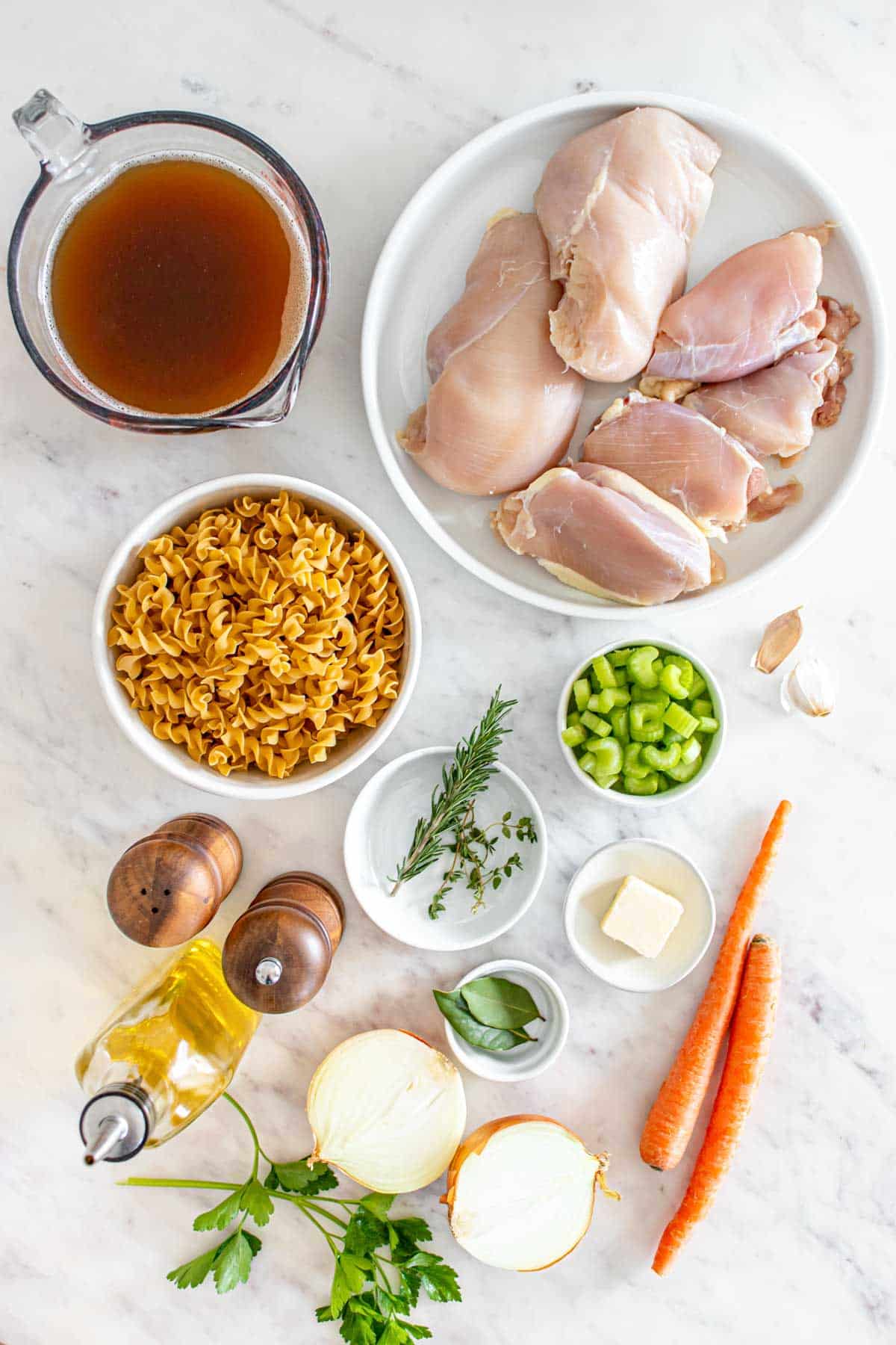 Ingredients for a chicken recipe laid out on a marble countertop, including raw chicken, pasta, vegetables, and broth.