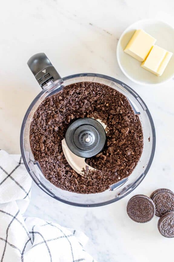 A food processor containing crushed chocolate cookies, with a plate of butter and whole cookies nearby on a marble surface.