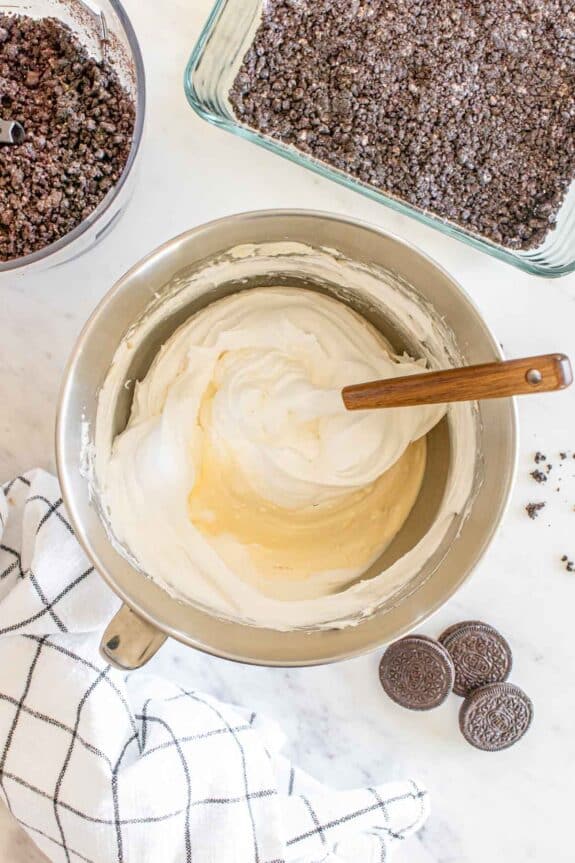 Mixing bowl with cream cheese frosting beside a bowl of crushed cookies and a dish of cookie crumble, with whole cookies and a kitchen towel on a marble countertop.