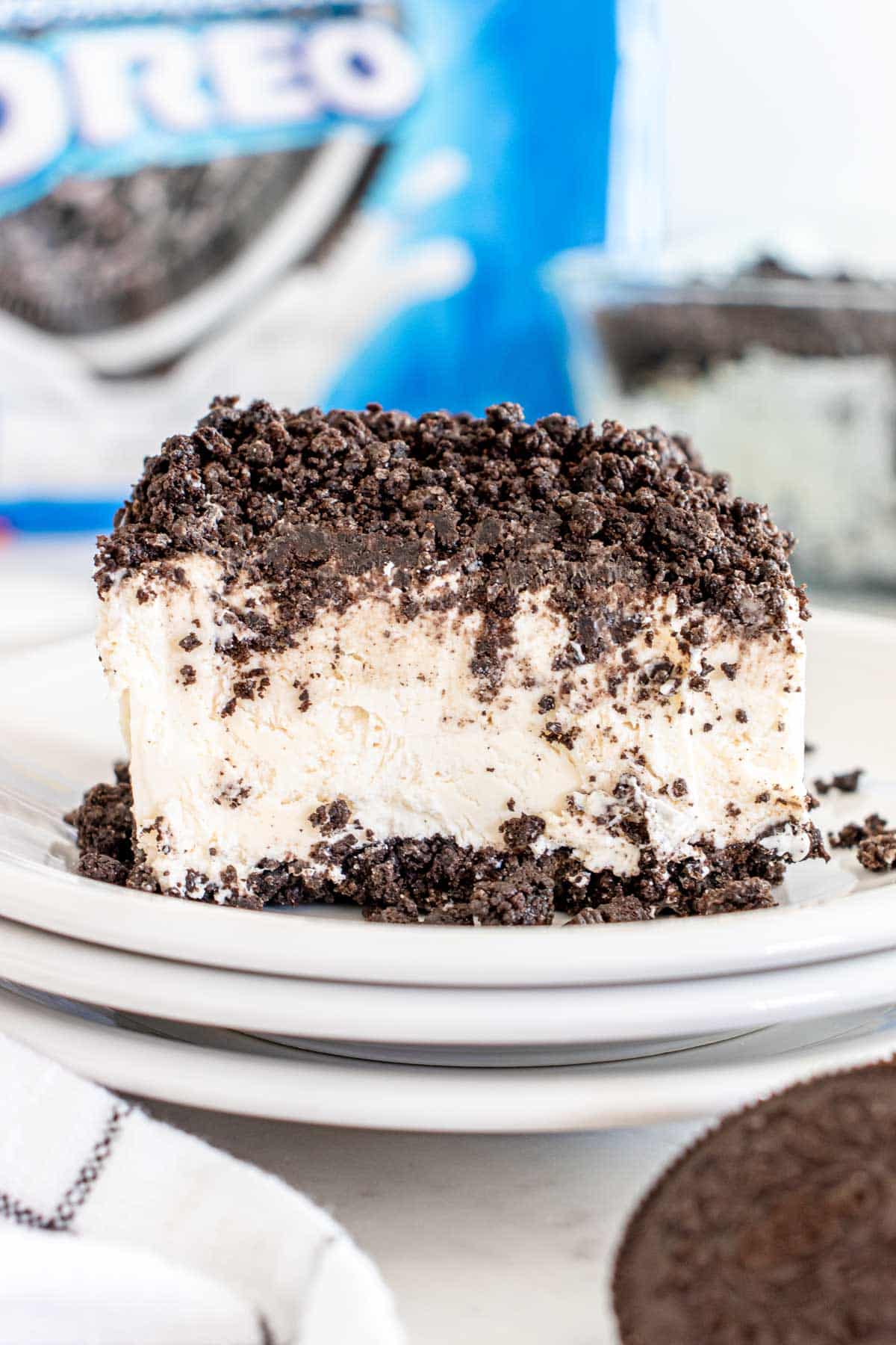 A slice of cookies and cream ice cream cake on a white plate, coated with crushed cookie crumbs, with a package of oreo cookies visible in the background.