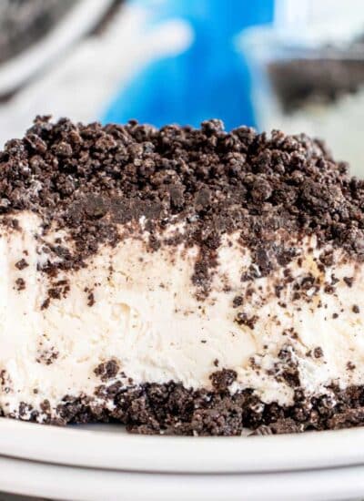 A slice of frozen cookies and cream ice cream cake on a white plate, topped with crumbled Oreo cookies.