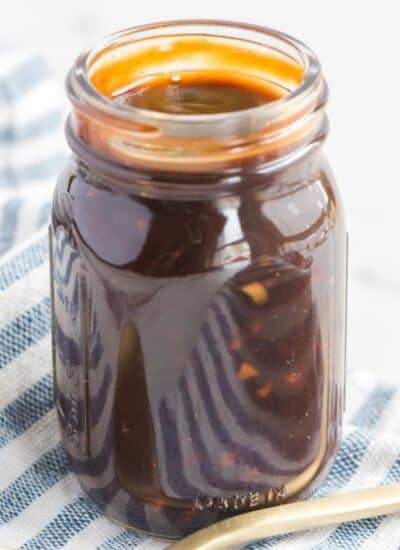 A glass jar filled with homemade teriyaki sauce on a blue and white checkered cloth with a gold spoon resting beside it.