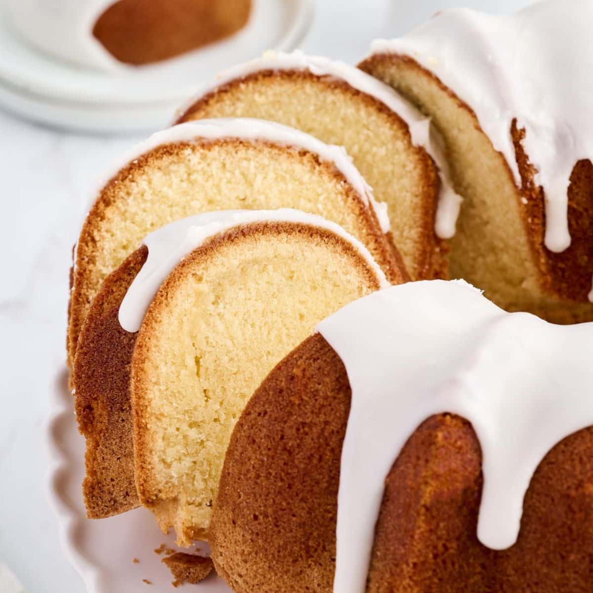 A sliced bundt cake with white icing is displayed on a white plate. A small plate with additional cake is in the background.