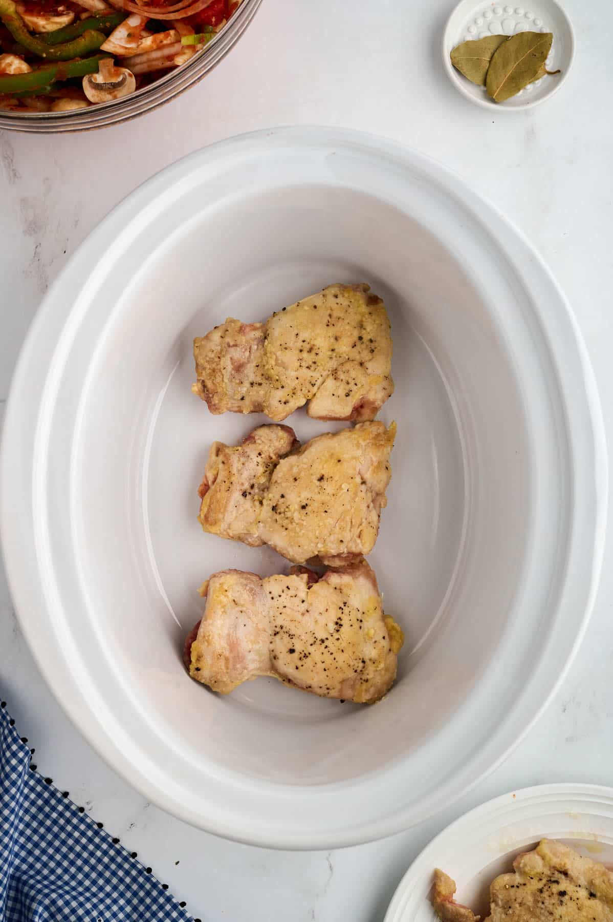 Three cooked chicken thighs are placed at the bottom of a white slow cooker. A small bowl with spices, a dish, and a blue checkered cloth are nearby.