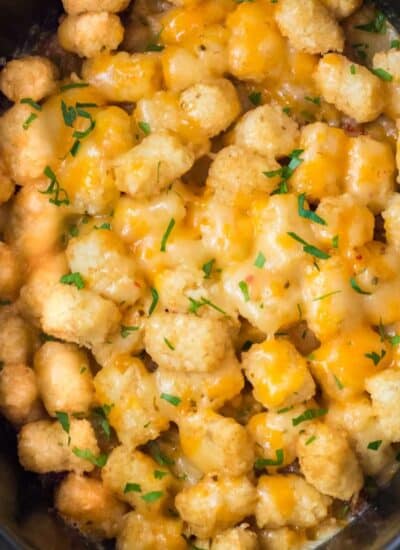 A close-up view of a slow cooker filled with tater tot casserole topped with melted cheddar cheese and sprinkled with chopped chives.